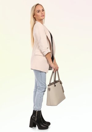 Frau mit langen blonden Haaren, die einen beigen Blazer, hellblaue Jeans, schwarze Absatzstiefel trägt und eine beige Handtasche mit beiden Händen hält.