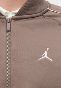 Gros plan d'une veste taupe zippée avec col côtelé et logo Jumpman blanc sur la poitrine gauche, au-dessus d'un col de chemise blanc.