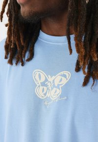 Camiseta de algodón azul claro con un gráfico de mariposa blanca y un diseño de texto en el pecho, con un escote redondo y mangas cortas.