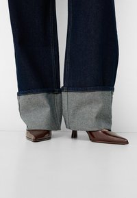Jeans de mezclilla azul oscuro de pierna ancha con un dobladillo gris claro, combinados con tacones de charol marrón de puntera afilada; costuras y texturas visibles.