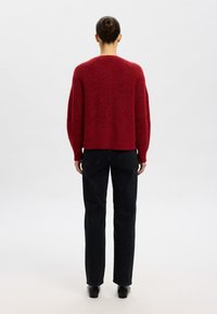 Raudonas megztas megztinis su laisvu siluetu, suapvalintu kaklu, nugara nuleistomis pečių linijomis ir ribotais rankogaliais. Dėvimas su tamsiais siaurėjančiais džinsais.