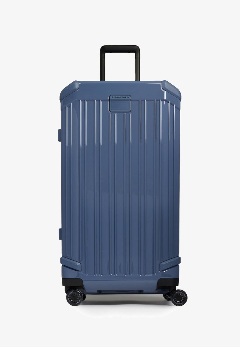 Valise rigide bleue à rainures verticales, poignée télescopique noire, quatre roues pivotantes noires, et petit panneau logo en haut.