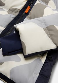 Polstret jakke med et camouflagemønster i grå, hvid og marineblå. Inkluderer en lynlåslukning med orange detaljer og elastiske manchetter.