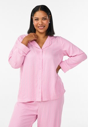 Lachende vrouw met halflang zwart haar die roze-wit gestreepte pyjama met knopen draagt, met de ene hand op haar heup en de andere bij haar kraag.