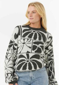 Donna bionda che indossa un maglione a maniche lunghe con motivo floreale in bianco e nero e jeans azzurri.