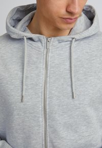 Sudadera gray con cremallera, acabado texturizado, cordones con puntas de metal y una tela suave. Presenta una capucha y una cremallera frontal.