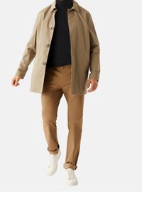 Beige leichter Mantel mit Knopfleiste, marineblaue Rollkragenjacke und hellbraune Hose. Weiße Sneaker runden das Outfit ab und betonen einen modernen Stil.