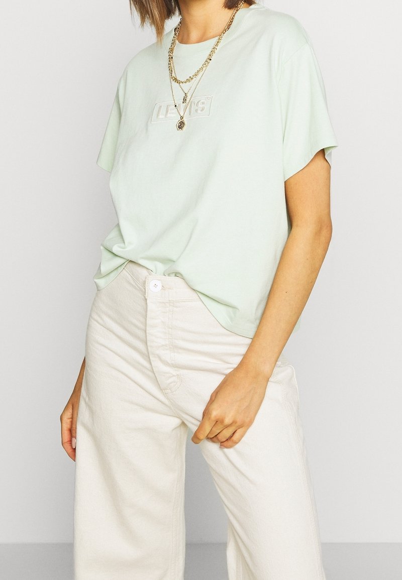 Femme portant un t-shirt Levi's vert clair rentré dans un pantalon large taille haute couleur écru, accessoirée avec des colliers dorés superposés.
