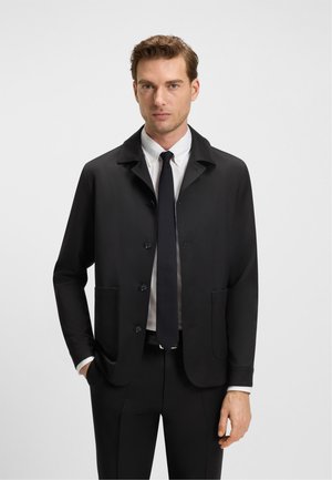 Giacca nera con colletto, cinque bottoni e due tasche frontali. Tessuto liscio, vestibilità sartoriale, indossata sopra una camicia bianca e una cravatta.