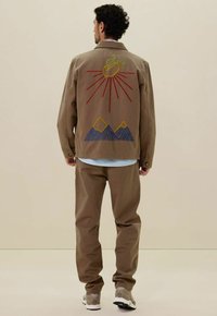 Bruine katoenen jas met rode en gele geborduurde zon, groene appel en blauwe bergpatronen; gecombineerd met bijpassende bruine broek.