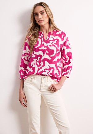 Kobieta o długich blond włosach, ubrana w różowo-białą bluzkę z abstrakcyjnym wzorem i kremowe spodnie, stoi z jedną ręką na biodrze.