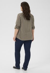 Woman with long wavy hair wearing a rolled-up striped shirt, dark blue jeans, and black shoes, standing facing away on a plain background.