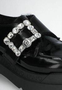 Black patent leather shoe featuring a large crystal-embellished buckle, thick black sole with textured accents, and soft lining.