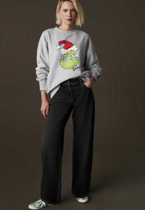 Grauer Kapuzenpullover mit einem bunten Grinch-Design, kombiniert mit hoch taillierten, weiten schwarzen Jeans und silbernen Sneakers.
