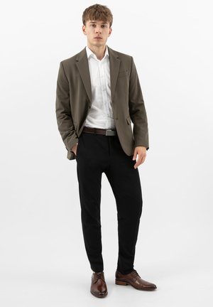 Jonge man staand, gekleed in een bruine blazer, wit overhemd, zwarte broek en bruine nette schoenen, tegen een witte achtergrond.