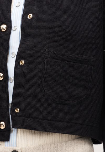Μαύρη πλεκτή ζακέτα με χρυσά κουμπιά πάνω σε ριγέ μπλε και λευκό πουκάμισο, με μπροστινή τσέπη και μπεζ ριμπ ύφασμα από κάτω.