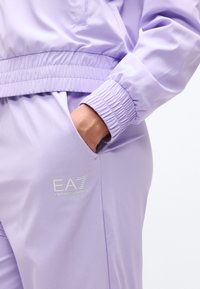 Χέρι ατόμου στην τσέπη ανοιχτόμωβ φόρμας Emporio Armani EA7 με ελαστικά μανσέτα και ζώνη μέσης ορατά.