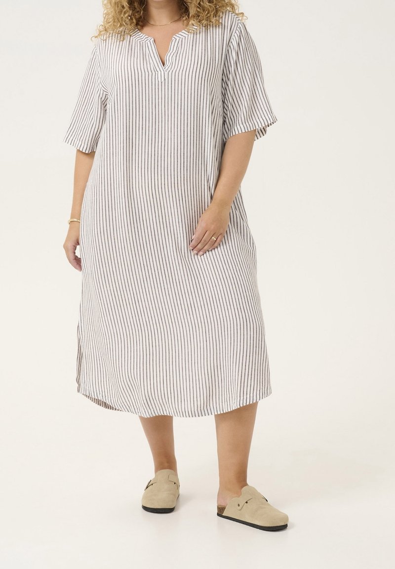 Femme portant une robe blanche ample jusqu'aux genoux avec de fines rayures verticales noires et des chaussures beiges sans lacets, debout sur un sol blanc.