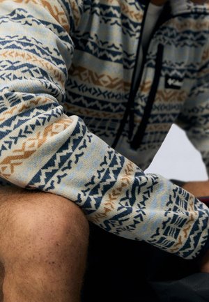 Persona che indossa una giacca Columbia con zip e motivo geometrico blu, beige e bianco, seduta con un ginocchio visibile.