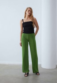 Haut noir sans bretelles avec des pois texturés, associé à un pantalon vert à taille haute et à jambes larges. Le mannequin porte des sandales noires sur un fond neutre.