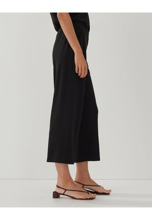 Kobieta stojąca, ubrana w czarne szerokie spodnie o długości do połowy łydki oraz czarne sandały na niskim obcasie z paskami, na jednolitym tle.