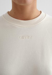 Felpa color crema con colletto a costine e dettaglio del logo in rilievo. Tessuto morbido, design minimalista, vista ravvicinata focalizzata sul torso superiore.