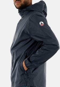 Veste coupe-vent navy avec capuche, poignets élastiques et poche latérale. Présente un logo circulaire sur la manche gauche. Matériau lisse et léger.