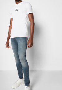 Bílé bavlněné tričko s malým černým logem na hrudi, doplněné o světle modré slim fit džíny a bílé tenisky. Hladká textura, jednoduchý design.