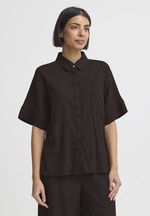 Woman with short dark hair wearing loose-fit dark brown short-sleeve button-up shirt and matching pants, standing against plain background.