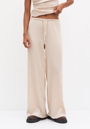 Frau trägt beige strukturierte, weit geschnittene Hosen mit Kordelzug in der Taille und braune klobige Schuhe, cropped Top teilweise sichtbar.