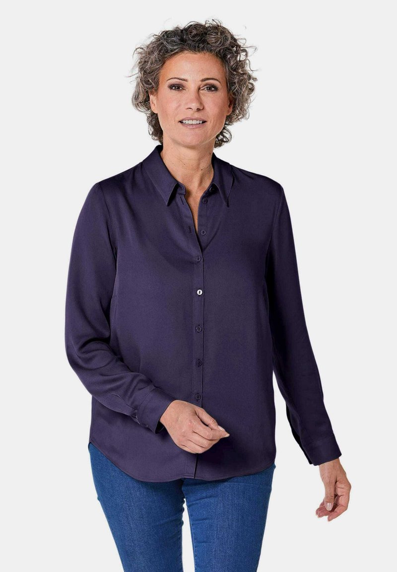 Long-sleeved navy blouse with a button-up front, collar, and curved hem. Smooth texture with a relaxed fit, paired with blue jeans.