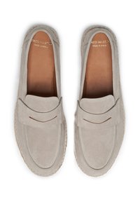 Mocassins en suède gris clair avec un accent cousu et une semelle en caoutchouc plate. L'intérieur présente une semelle brune avec le marquage de la marque. Fabriqué en Espagne.