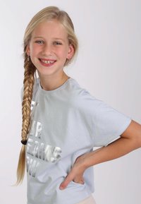 Lichtblauwe katoenen t-shirt met korte mouwen met zilveren tekst "HAAL JE DROMEN NU." Persoon heeft lang, gevlochten haar en ringoorbellen.