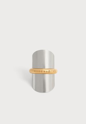Bague de couleur dorée avec des chiffres gravés de 0 à 9, le chiffre 5 entouré, présentée sur un support cylindrique métallique sur fond blanc.
