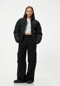 Chaqueta acolchada negra con cuello alto, textura acolchada, botones de presión, combinada con pantalones cargo negros y sueltos. Calzado robusto completa el conjunto.