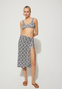 Femme portant un haut de bikini à motifs noir et blanc et une jupe enveloppante assortie avec une fente sur le côté, accessoirisée avec des boucles d'oreilles créoles et des sandales marron.