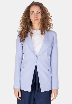 Donna con capelli ricci che indossa un blazer azzurro chiaro sopra una camicia bianca e pantaloni scuri, in piedi davanti a uno sfondo bianco uniforme.