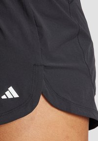 Melni sporta šorti ar izliektu apakšmalu, gludu tekstūru un baltu Adidas logo kreisajā apakšējā stūrī.