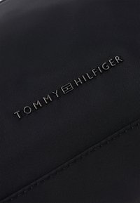 Crni najlon s teksturiranom površinom, s utisnutim logotipom "TOMMY HILFIGER", uz prošivenu dekoraciju sa strane.