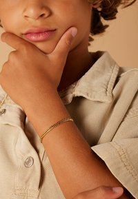 Gouden kettingarmband gedragen op een gebruinde arm, met een gestructureerd ontwerp. Beige overhemd met kraag en een gedeeltelijke knoopdetail zichtbaar op de achtergrond.