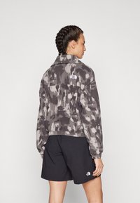 Giacca in fleece con motivo camouflage grigio, colletto e orlo elastico. Logo visibile sul retro. Abbinata a pantaloni corti neri.