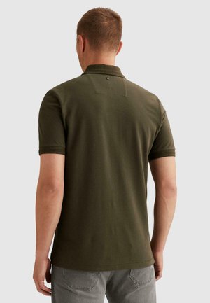 Man gezien van achteren, gekleed in een olijfgroen poloshirt met korte mouwen en grijze spijkerbroek, tegen een effen lichte achtergrond.