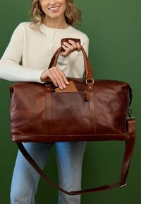 Rjava usnjena torba za potovanja z gladko teksturo, dvojni ročaji, odstranljiv ramenski trak in žep z zadrgo, ki vsebuje držalo za kartice.