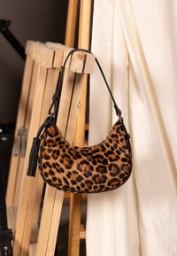 Sac à main imprimé léopard avec une forme courbée, une texture en fourrure douce, une bandoulière en cuir noir et un détail de pompon, suspendu devant un arrière-plan neutre.