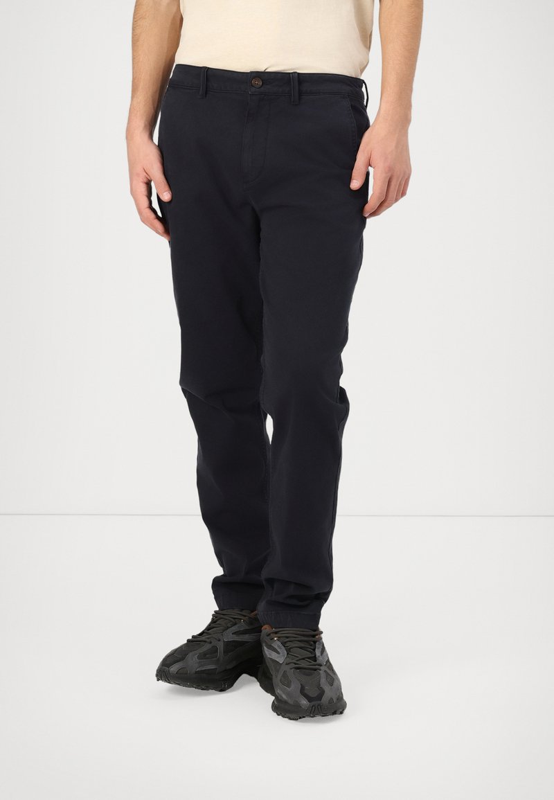 Marineblauwe chino's met rechte pijpen, gemaakt van katoen, met een knoopsluiting, voorzakken en een nette afwerking. Draagt met zwarte sportschoenen.