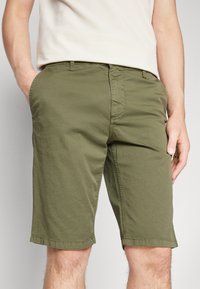Pantaloni corti verde oliva con taglio dritto, realizzati in tessuto di cotone. Presentano tasche frontali, chiusura con bottone e orli cuciti.