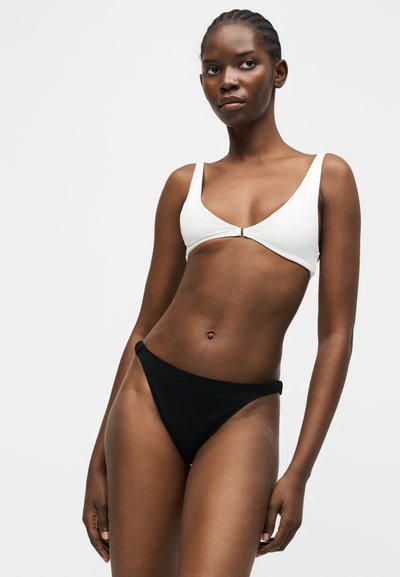 Νέα γυναίκα με πλεξούδες, που φοράει λευκό τοπ μπικίνι με κλιπ μπροστά και μαύρο κάτω μέρος μπικίνι, με φόντο απλό.