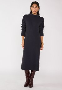 Abito a maglia blu navy con collo alto e maniche lunghe, caratterizzato da polsini e orlo a coste. Indossato con stivali a ginocchio lucidi color bordeaux.
