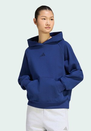 Ung kvinde iført en blå hættetrøje med hænderne i forlommen og lysegrå joggingbukser, der kigger mod højre.