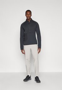 Fekete negyed cipzáras pulóver, texturált mintával és elülső zsebbel, világosszürke nadrággal és fekete sportcipővel párosítva.